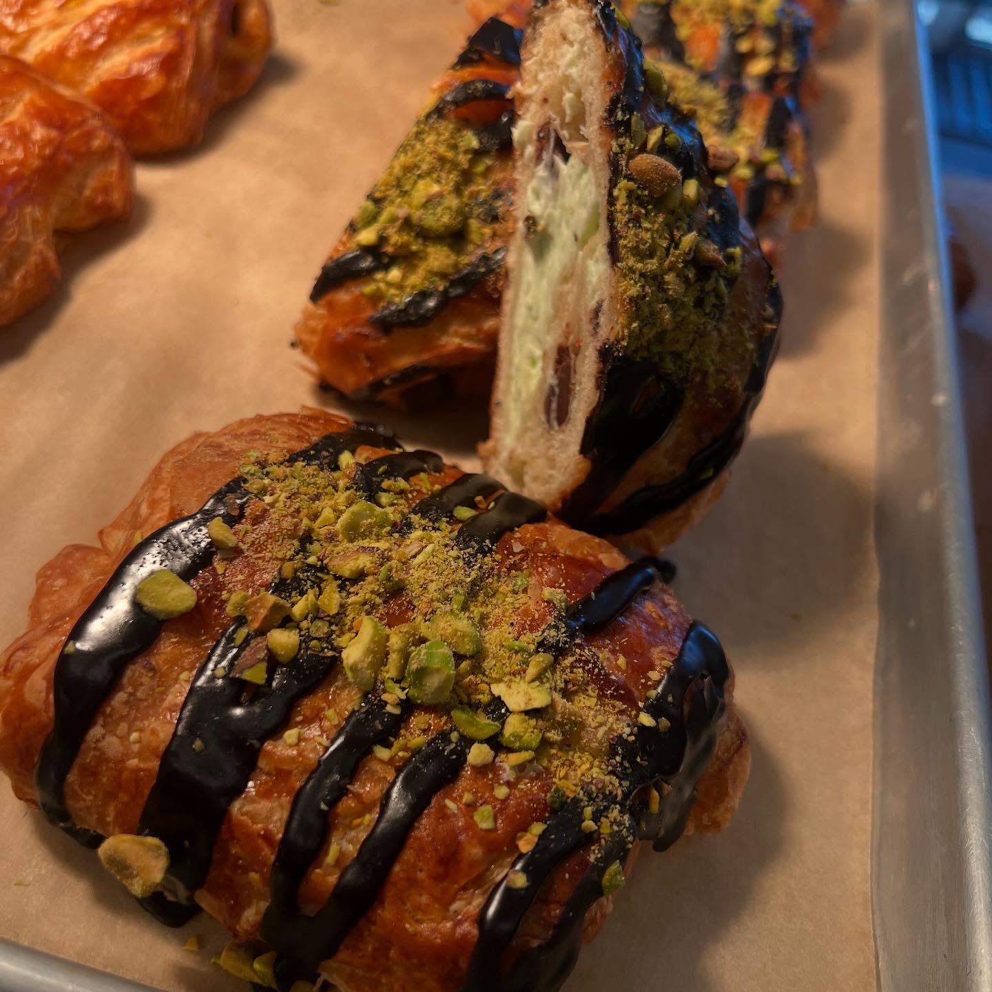 Homemade pistachio cream-filled croissants drizzled with melted chocolate and topped with crushed pistachios, displayed whole alongside a cross-section view of the flaky pastry and creamy filling